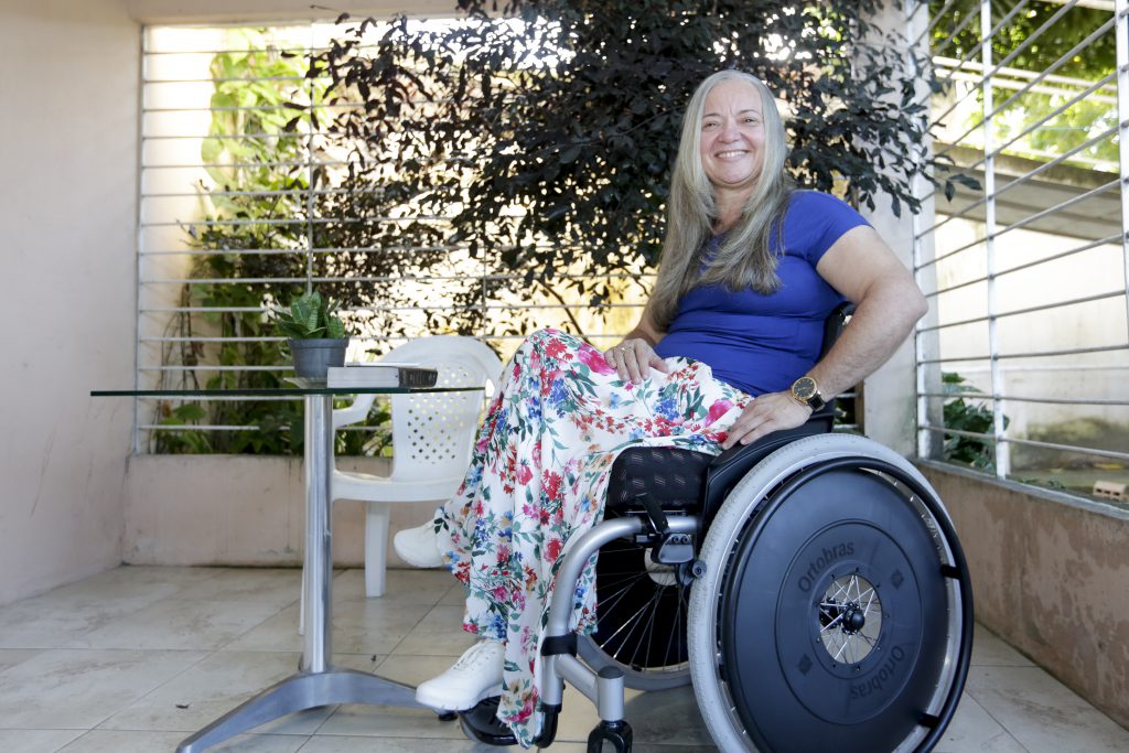 Na foto, uma mulher cadeirante sorri no terraço de casa.