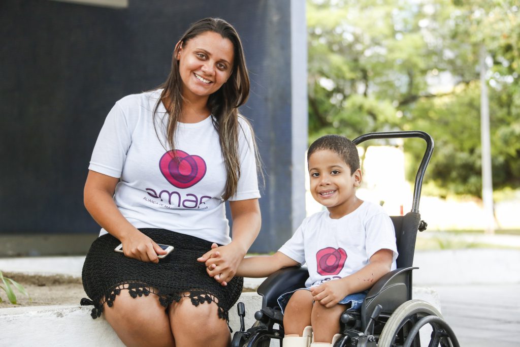 Foto de uma mulher sentada ao lado de uma criança que está na cadeira de rodas. Os dois sorrem.