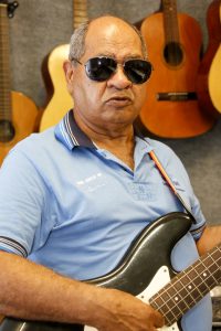 Foto em sala com violões pendurados na parede. Homem com óculos escuros segura uma guitarra.
