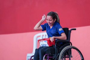 Na foto, menina vestida de uniforme de atleta e em cadeira de rodas comemora com um dos braços erguidos.