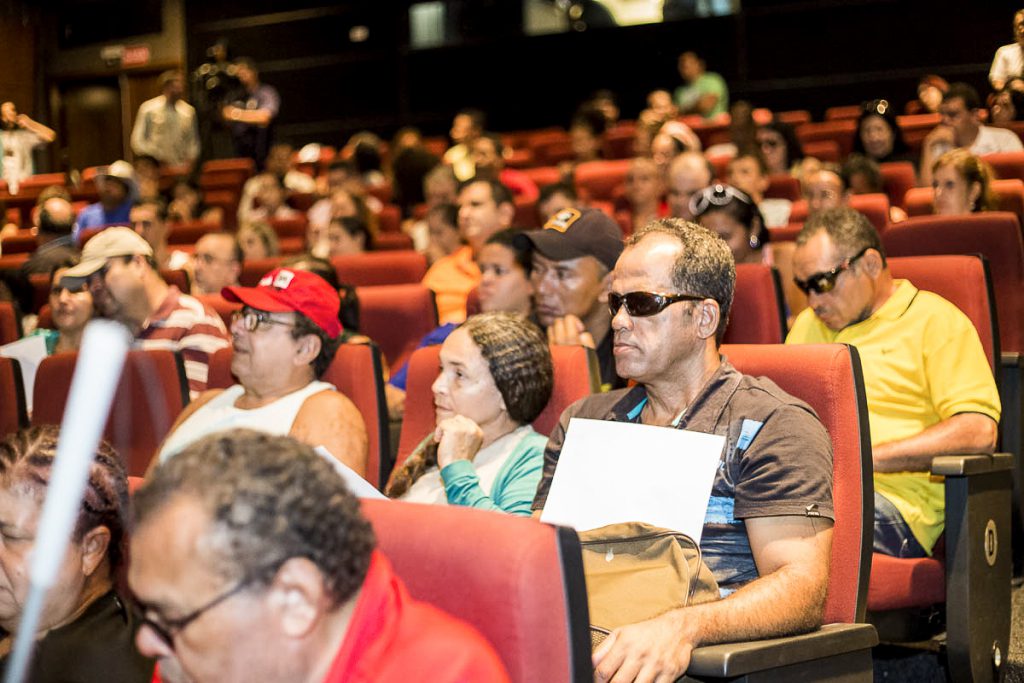 Na foto, várias pessoas sentadas numa sala de cinema com homem de óculos escuros em primeiro plano.