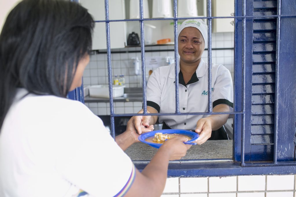 Foto de merendeira entregando refeição a aluna de escola pública.