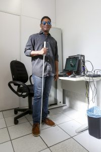Foto do jornalista Vinícius Passos em pé no local de trabalho dele, com uma bengala na mão esquerda.
