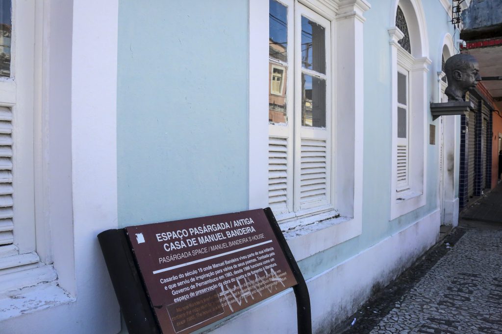 Foto da fachada do Espaço Pasárgada, um sobrado verde claro de janelas brancas na Rua da União. Em destaque, uma placa que informa a importância histórica da casa onde Manuel Bandeira viveu na infância. Ao fundo, o busto do poeta que fica na entrada do prédio.