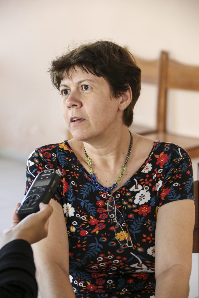 Foto da professora Maria Bethânia sendo entrevistada em uma sala, mostrando a mão da repórter em primeiro plano.