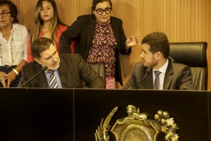 Foto do deputado Tony Gel falando ao microfone com o deputado Joaquim Lira ao lado, ambos sentados em sala do Plenarinho.