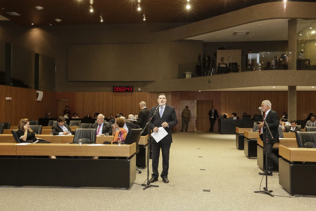 Foto do deputado Tony Gel falando ao microfone de aparte no centro do Plenário, com demais deputados sentados nas cadeiras ao fundo.