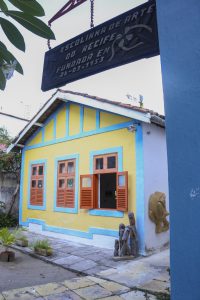 Foto de casa onde funciona a Escolinha de Arte do Recife. Casa é amarela com detalhes verdes e janelas marrons, possui quintal na frente.
