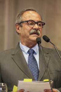 Foto do secretário Pedro Eurico com manual nas mãos, falando ao microfone da tribuna da Alepe.