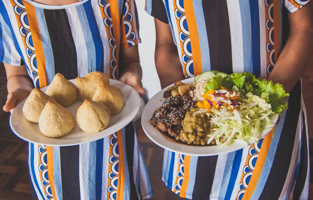 Foto mostra tronco de duas pessoas que seguram pratos com alimentos veganos: coxinhas e alimentos diversos.