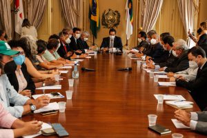 Governador aparece ao fundo de uma mesa longa no Palácio, com brasão do Estado e bandeiras do Brasil e de Pernambuco atrás dele. Estão sentados, dos dois lados da mesa, parlamentares e lideranças de trabalhadores rurais.