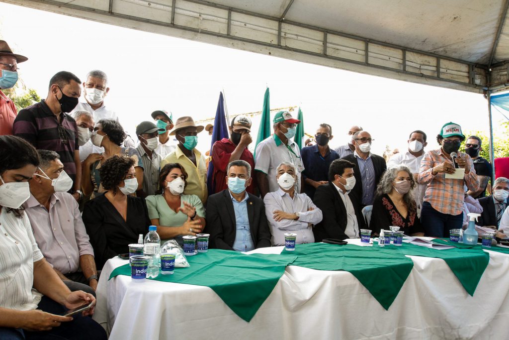 Mesa em local aberto, coberta com um toldo, com parlamentares sentados e rodeada por trabalhadores rurais