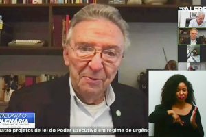 Deputado fala em reunião virtual, com mesa de escritório e estante de livros ao fundo