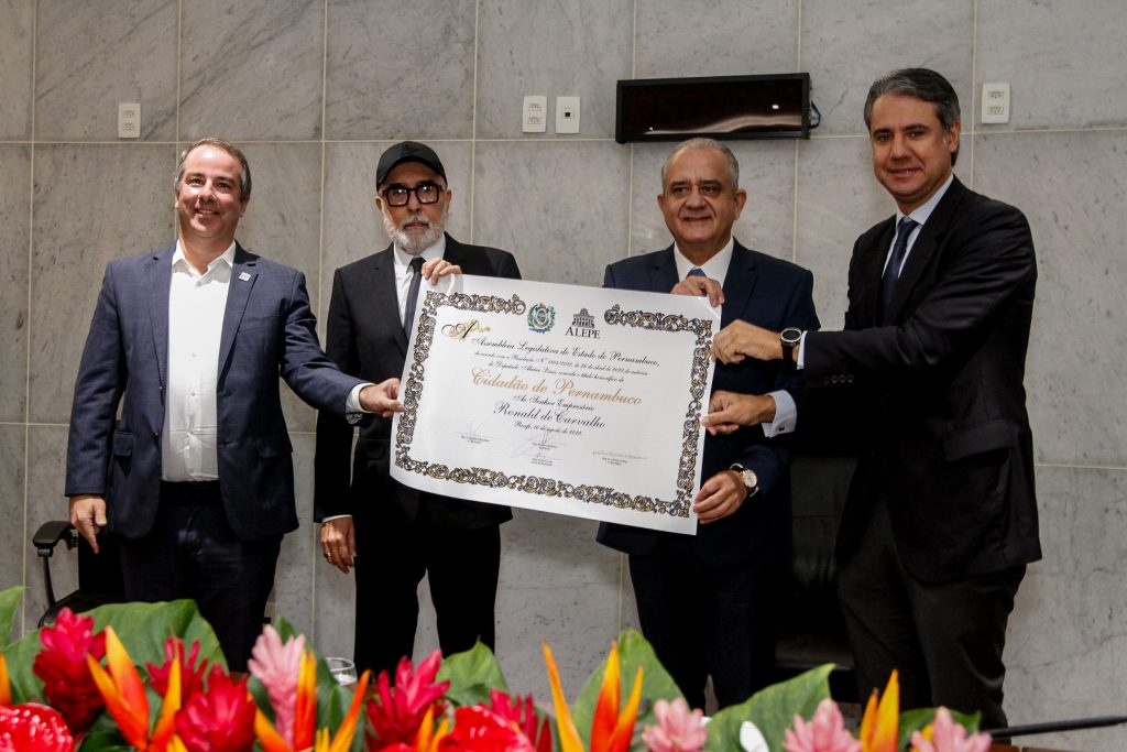 Empresário Ronald de Araújo, de boné, segura diploma em tamanho grande (A2). Ele tem barbas grisalhas, quase brancas, e usa óculos quadrados. Ao lado dele, está o deputado Aluísio Lessa. Junto com eles, outras duas pessoas também de terno seguram o diploma