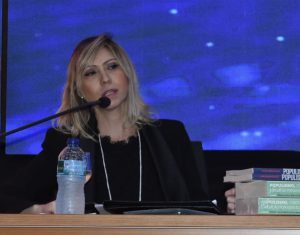 Mulher loira, com roupas sociais pretas, fala ao microfone em uma mesa de auditório, com painel azul ao fundo e uma garrafa de água mineral e livros grossos à frente