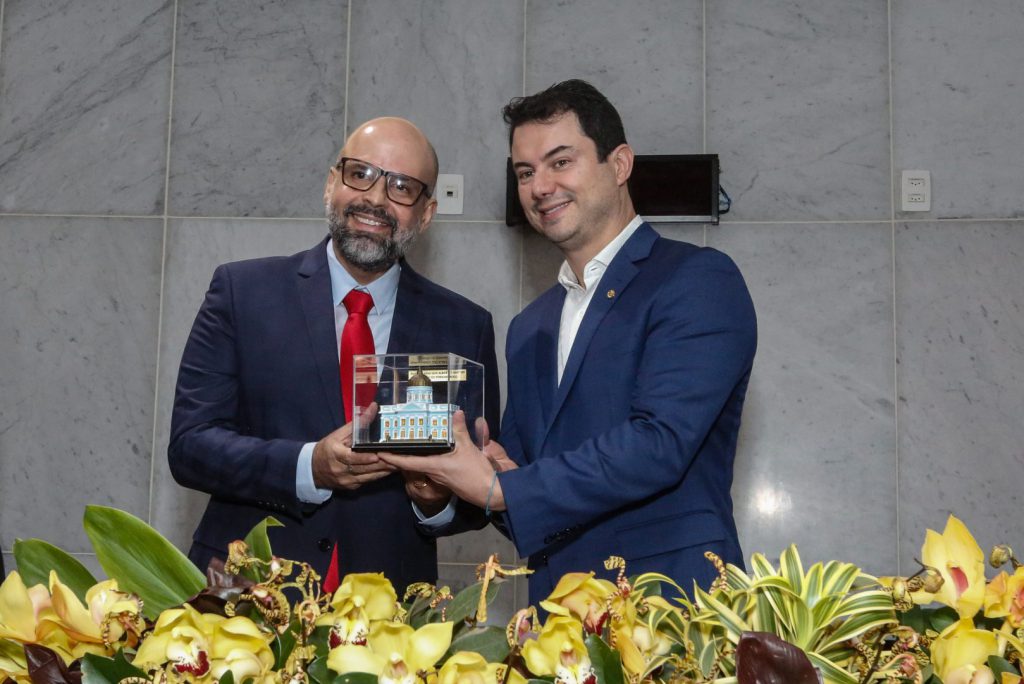 Médio, à direita, recebe réplica do Palácio Joaquim Nabuco do deputado, à direita. Todos estão de terno, e há um arranjo de flores à frente da mesa.