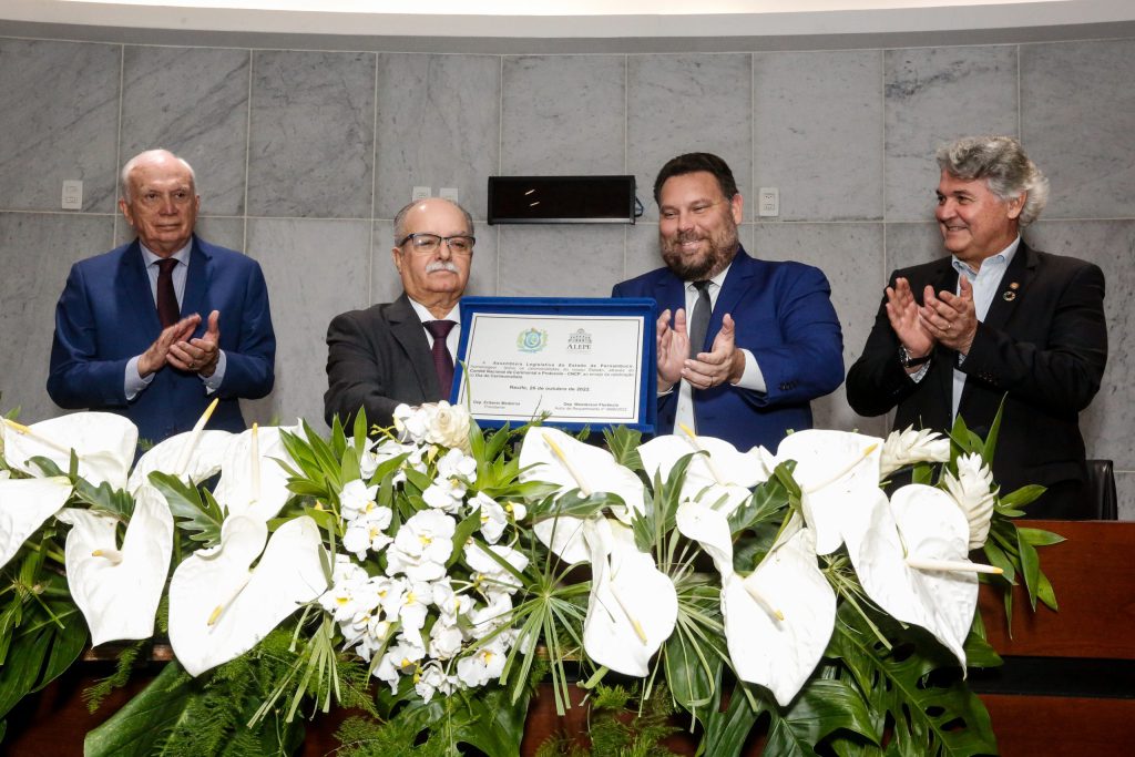 4 homens de terno em frente a uma placa de homenagem, numa mesa com arranjo de flores brancas. O terceiro, da esquerda para direita é o deputado Wanderson Florêncio.