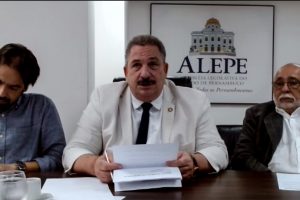 Deputados Diogo Moraes, Eriberto Medeiros e Francismar Pontes sentados numa mesa com papéis um copo de água à esquerda. Eriberto, vestindo terno branco e gravata preta, segura alguns papéis. Logotipo da Alepe está na parede ao fundo.