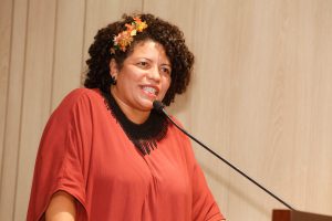 Deputada, com roupas vermelhas em tons terrosos, fala ao microfone do auditório Sérgio Guerra