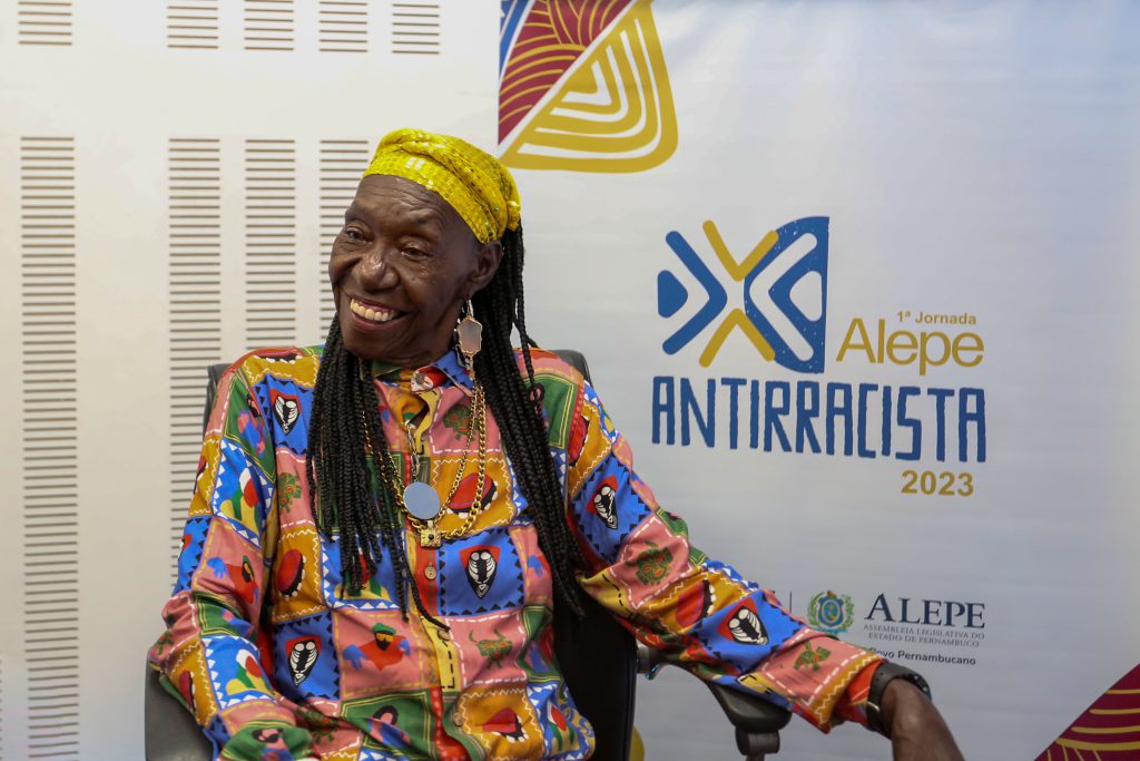 Foto de Lia de Itamaracá, com roupas coloridas. Atrás dela, adesivação com logotipo da Jornada Alepe Antirracista