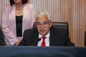 Deputado Antonio Moraes, usando terno e gravata vermelha, sentado ao microfone. Corpo de assessora, usando tailleur rosa, aparece atrás dele.