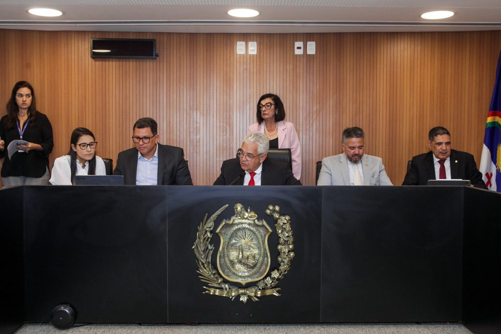 Foto do Plenarinho da Alepe, com deputado Antonio Moraes ao microfone. Assessores e deputados sentados atrás de uma bancada preta, com o brasão de armas de Pernambuco à frente. Duas assessoras estão em pé, atrás deles, à frente de um painel de madeira.