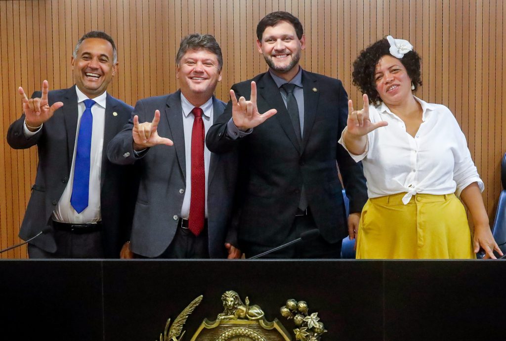 Deputados Nino de Enoque, Sileno Guedes, Gilmar Júnior e Dani Portela sorriem e fazem o sinal de Libras para "eu te amo". Eles estão em pé, atrás da mesa da sala de reuniões da comissão.
