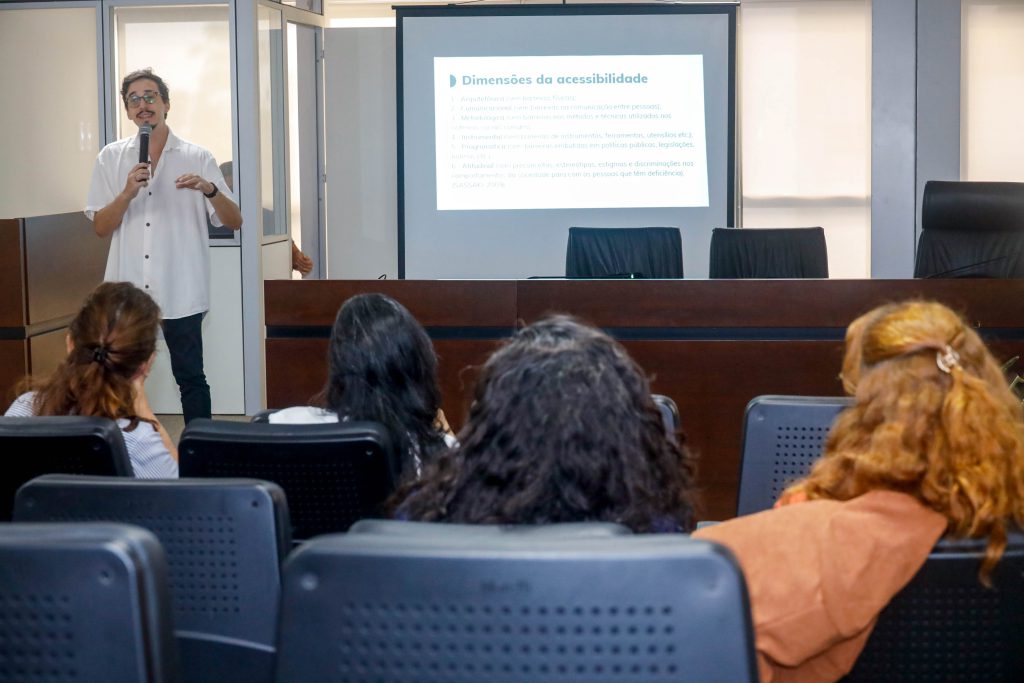 Professor Túlio Rodrigues fala em microfone para turma de alunos, que estão de costas. Ele é um homem alto, magro, veste uma camisa branca e usa óculos. O local é um auditório com paredes brancas e móveis em madeira escura. Ao fundo, parte de uma tela com a projeção do conteúdo da aula.
