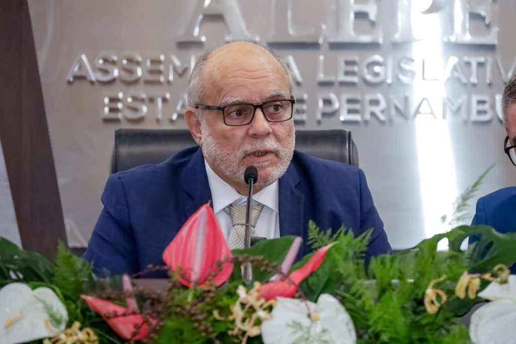 Foto do deputado falando ao microfone. Ele tem barba branca curta, usa óculos, terno azul com gravata bege clara. A frente dele, um arranjo de flores, e atrás, painel com o nome "Alepe - Assembleia Legislativa do Estado de Pernambuco" em letras de alumínio na parede.