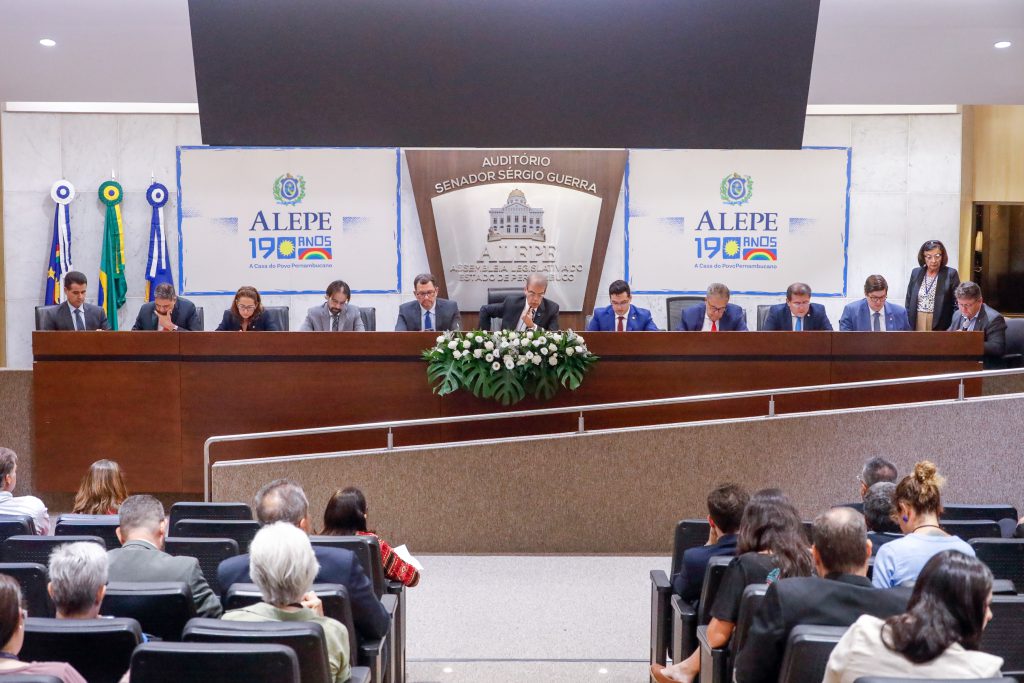 Mesa do auditório Sérgio Guerra, com todas as cadeiras preenchidas por deputados estaduais usando terno e gravata. Atrás dos deputados, do lado esquerdo, estão as bandeiras de Pernambuco, do Brasil e do Recife, penduradas na parede. Ao centro, o nome "Auditório Sérgio Guerra" e um emblema da Alepe com o Palácio Joaquim Nabuco estilizado. Dos dois lados de emblema, o logotipo "Alepe 190 Anos". À frente da mesa onde estão os deputados, as primeiras fileiras do auditórios, parcialmente preenchidas pelo público. 