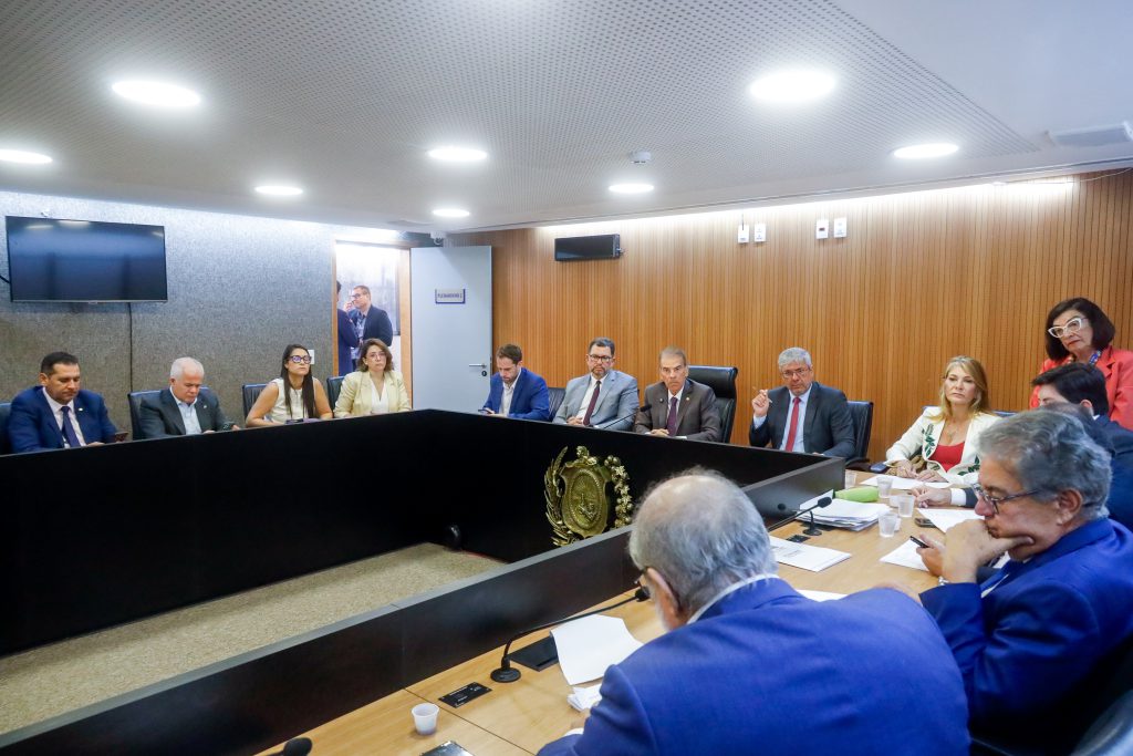 Mesa do Plenarinho da Alepe, vista em formato diagonal. A mesa em composto por deputados e secretários do governo estadual, enquanto assessores estão em pé e na porta do Plenarinho aberta, ao fundo. 