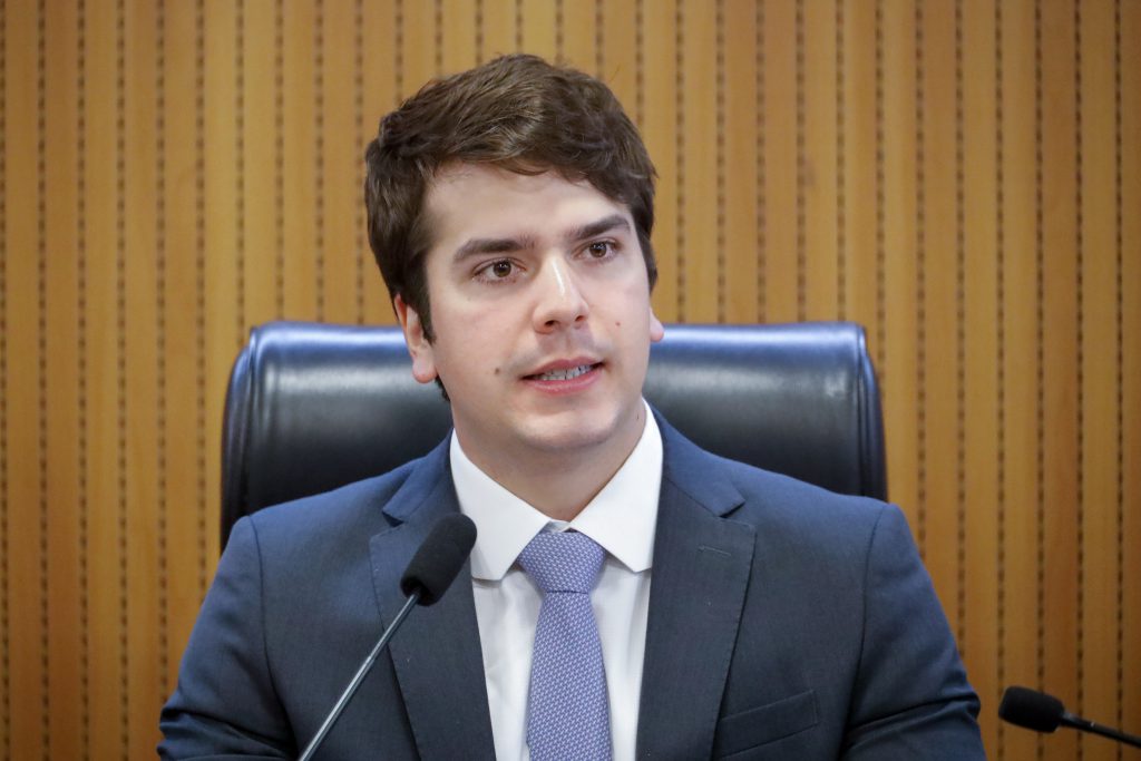 Deputado Antonio Coelho, com terno grafte e gravata em roxo claro, fala ao microfone sentado numa cadeira de couro. Ao fundo, um painel de madeira.