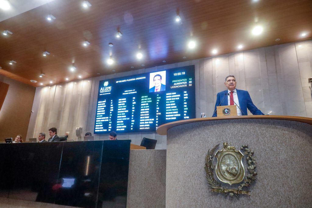 Deputado João Paulo, usando terno azul escuro e gravata vermelha, fala na tribuna da Alepe, com o painel e a Mesa Diretora ao fundo da foto. 