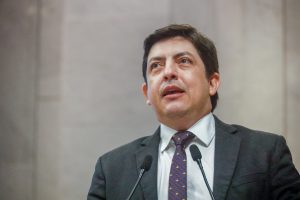 Deputado, um homem branco de cabelos pretos, lisos e curtos, de terno, fala ao microfone da tribuna. 