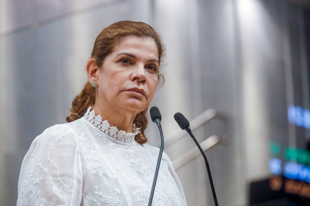 A deputada Socorro Pimentel, uma mulher branca com cabelos longos e presos, usando um vestido branco com golas de renda, fala ao microfone da tribuna. 