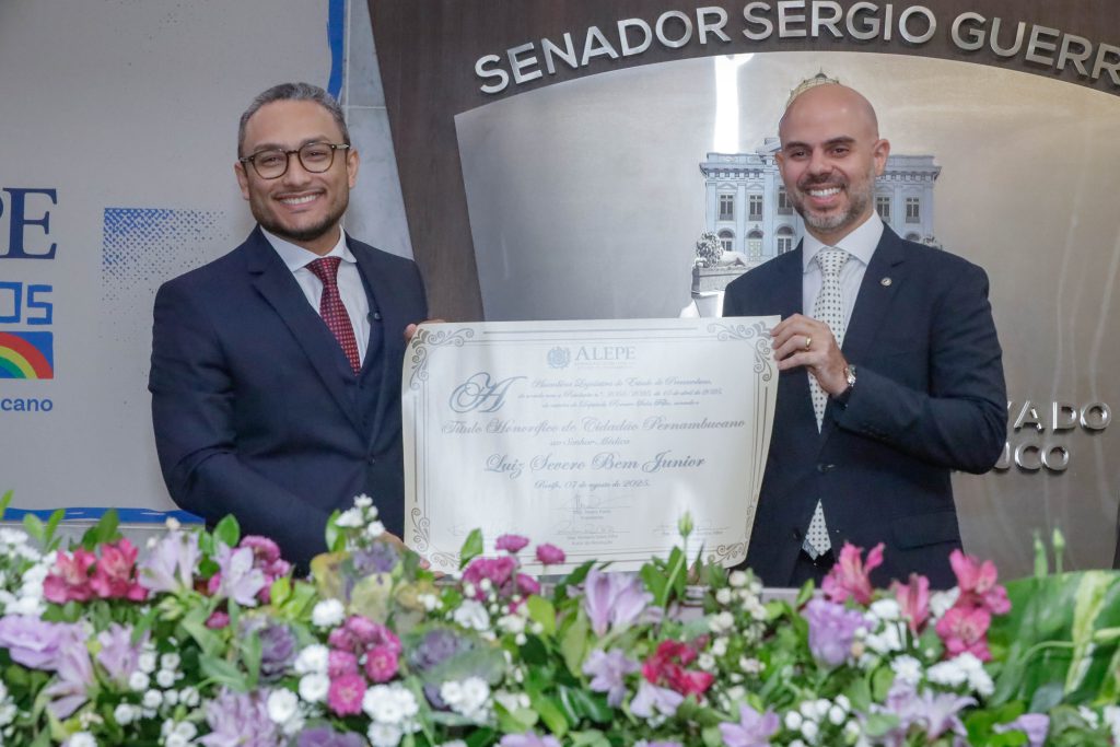 Médico Luiz Severo, usando terno azul marinho e gravata vermleha, óculos e cabelos e barba curtos, está a esquerda, segurando um diploma em formato grande, de cartaz. Do lado direito, segurando o mesmo diploma, está o deputado Romero Sales Filho, que usa terno azul marinho e gravata branca com detalhes pretos. O deputado tem o cabelo raspado e barba curta e grisalha. 