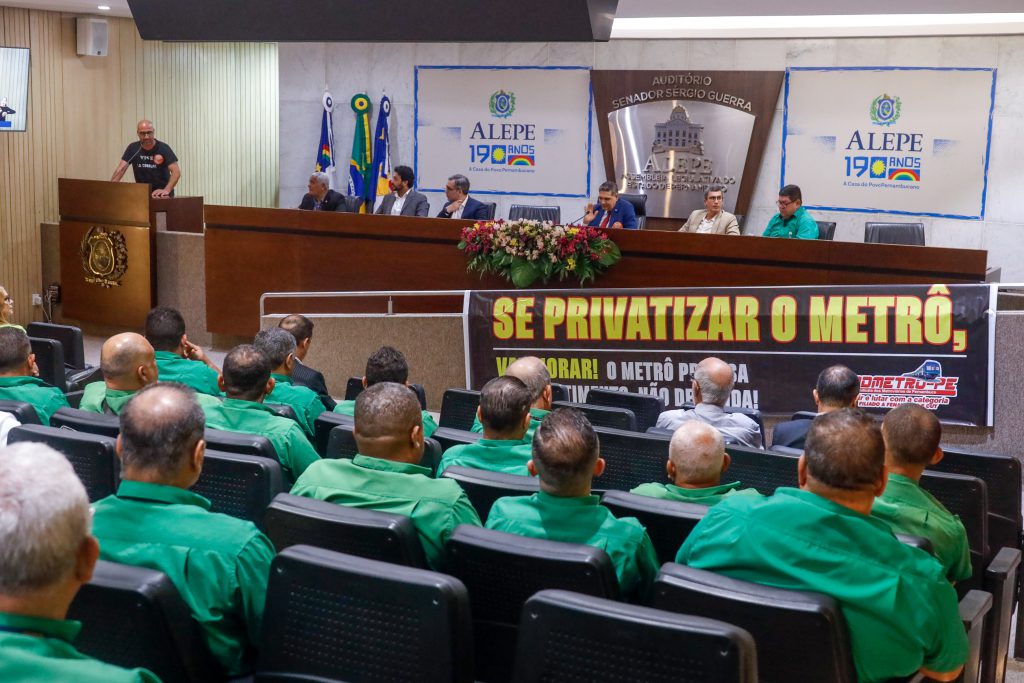 À frente, é possível ver as costas das pessoas assistindo a audiência pública, todas elas com camisas verde claro. Atrás, se vê a mesa do Auditório com autoridades e uma pessoa discursando. Abaixo da mesa, está uma faixa na cor preta com letras amarelas com o dizer "Se Privatizar o Metrô, vai piorar". Atrás da mesa, símbolos da Alepe na parede. 