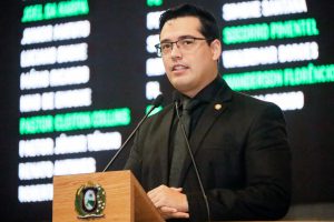 Deputado fala na tribuna, usando terno e gravata pretos. Atrás dele, nomes de parlamentares no painel eletrônico