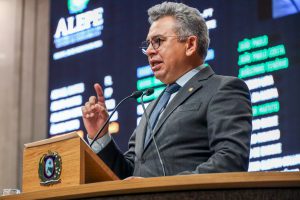 Deputado Doriel Barros, com terno cinza, fala na tribuna. Ao fundo, imagem desfocada do painel da Alepe