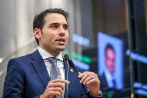 Deputado João Paulo Costa, com terno azul marinho e gravata cinza, fala na tribuna. Ao fundo, imagem desfocada do painel da Alepe, com foto do deputado