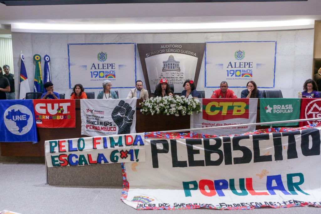 Mesa do auditório da Alepe, com a deputada Rosa Amorim usando boné do MST e a deputada Dani Portela ao centro. Na mesa, bandeiras de UNE, MST, Sindicato dos Bancários, CTB, Brasil Popular e MTST. Na rampa de acesso à mesa, bandeira com letras coloridas com os dizeres "Pelo fim da Escala 6x1" e "Plebiscito Popular". 