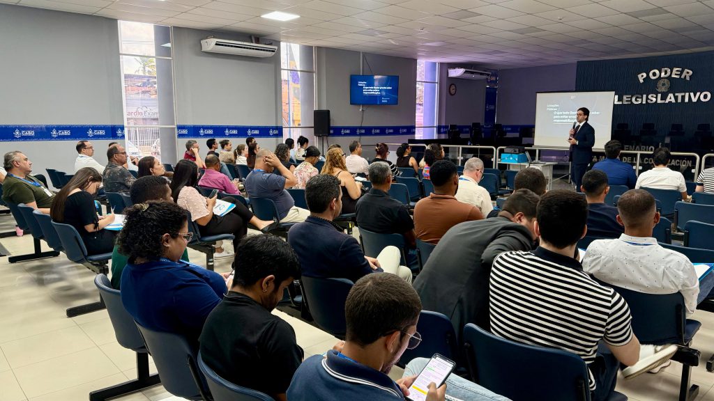 Sala do Plenário da Camara de Vereadores, com pessoas de costas sentadas nas fileiras de cadeiras. A sala é ampla, com paredes branco gelo, e na parede lateral há algumas faixas verticais de vidro levemente fosco que revelam detalhas da rua. Ao fundo, um quadro branco enquanto um professor fala, e uma TV na parede lateral. 