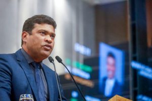 Deputado Joel da Harpa fala no microfone da tribuna, com reflexo da tela do painel eletrônico com sua foto ao fundo. Ele veste um terno azul escuro com camisa cinza e gravata da mesma cor do terno. 