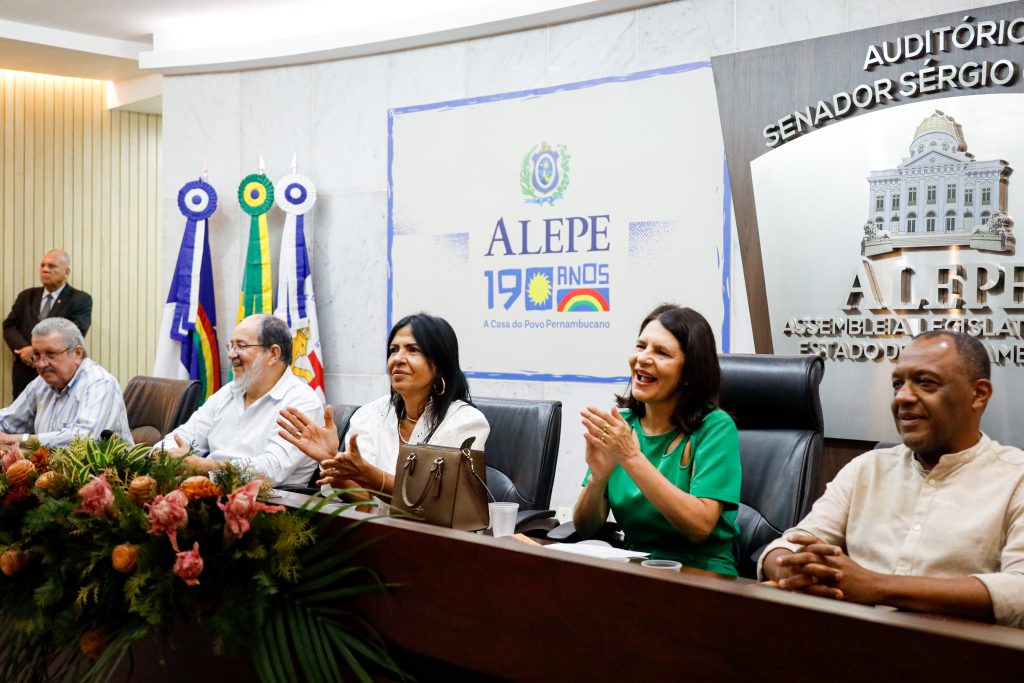 Mesa do auditório da Alepe, com três homens e duas mulheres sentados. As mulheres, sentadas ao meio, batem palma. Por trás deles, na parede, as bandeiras de Pernambuco e do Brasil à esquerda e o logotipo da Alepe ao centro. 