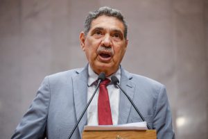 Deputado João Paulo, com terno cinza claro e gravata vermelha, fala no microfone da tribuna da Alepe. 