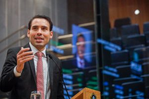 Deputado João Paulo Costa, usando terno preto e gravata vermelha. Ao fundo, à direita, aparece o painel do Plenário refletido no vidro da galeria
