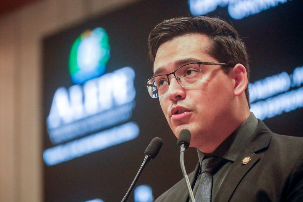 Foto em close do deputado Cayo Albino falando ao microfone, usando terno preto com gravata escura. Ao fundo à esquerda, desfocado, aparece o logo da Alepe no painel eletrônico do Plenário. 
