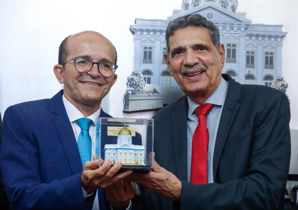 À esquerda, está o pastor Rinadlo Ce´zar, um homem brnaco, calvo, usando óculos e terno com gravata azul clara brilhante. Ele segura uma miniatura da Assembleia Legislativa junto com o deputado João Paulo, que está à sua direita, utilizando terno com gravata vermelho vivo. Ambos estão sorrindo. Ao fundo, por trás do deputado João Paulo, está um painel com uma reprodução do Palácio Joaquim Nabuco. 