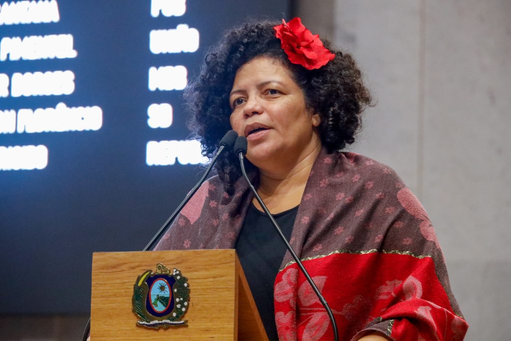 Dani Portela fala na tribuna, utilizando um Xale com a parte superior vinho e a de baixo em vermelho, um uma rosa vermelha como adorno do lado esquerdo do cabelo. 