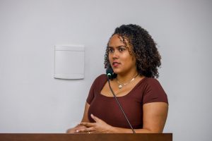 Morgana Xavier, uma mulher negra, de cabelos encaracolados e blusa marrom, fala no púlpito do auditório.